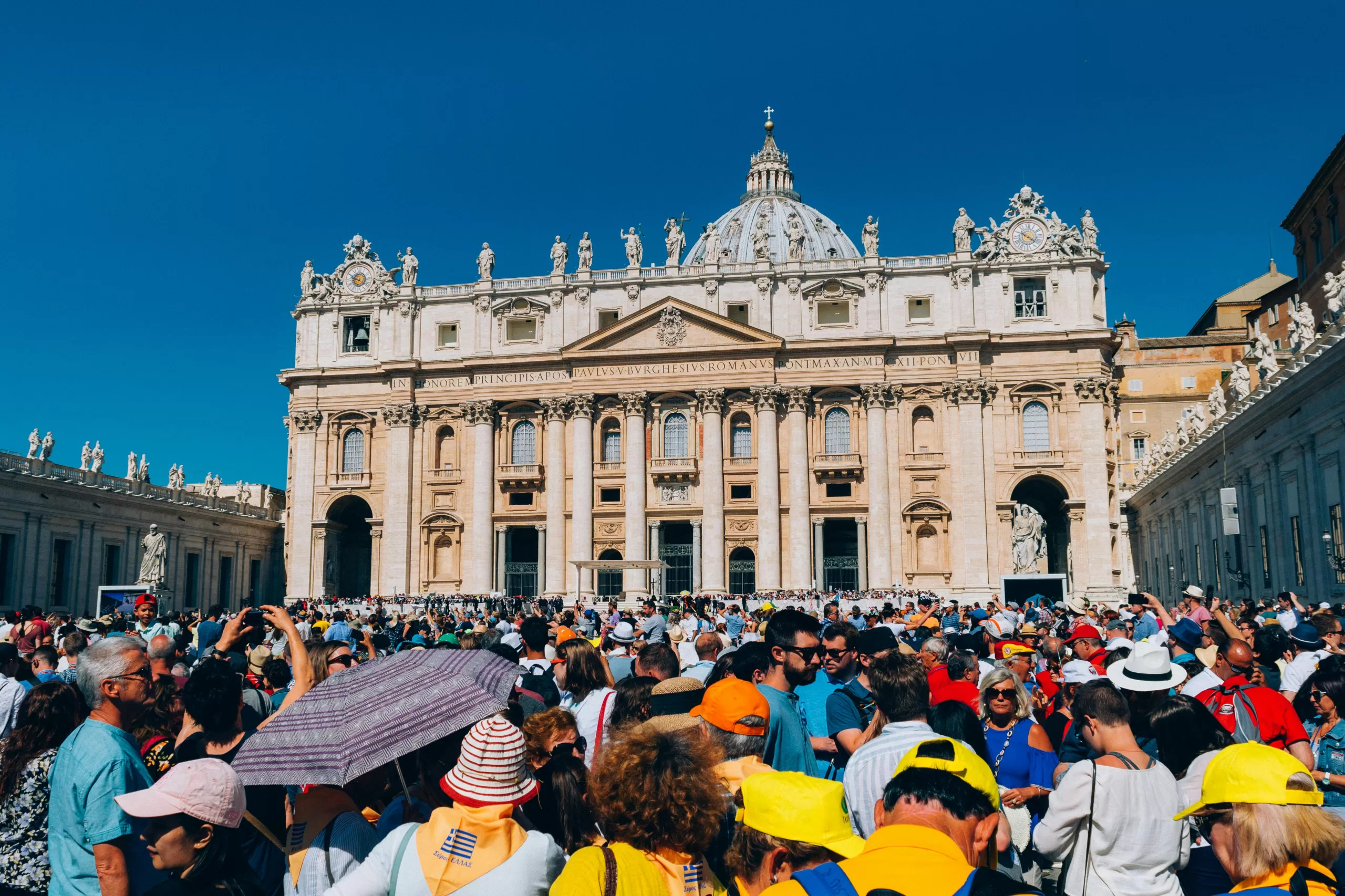 A large crowd gathers outside St. Peter's Basilica under a clear blue sky.